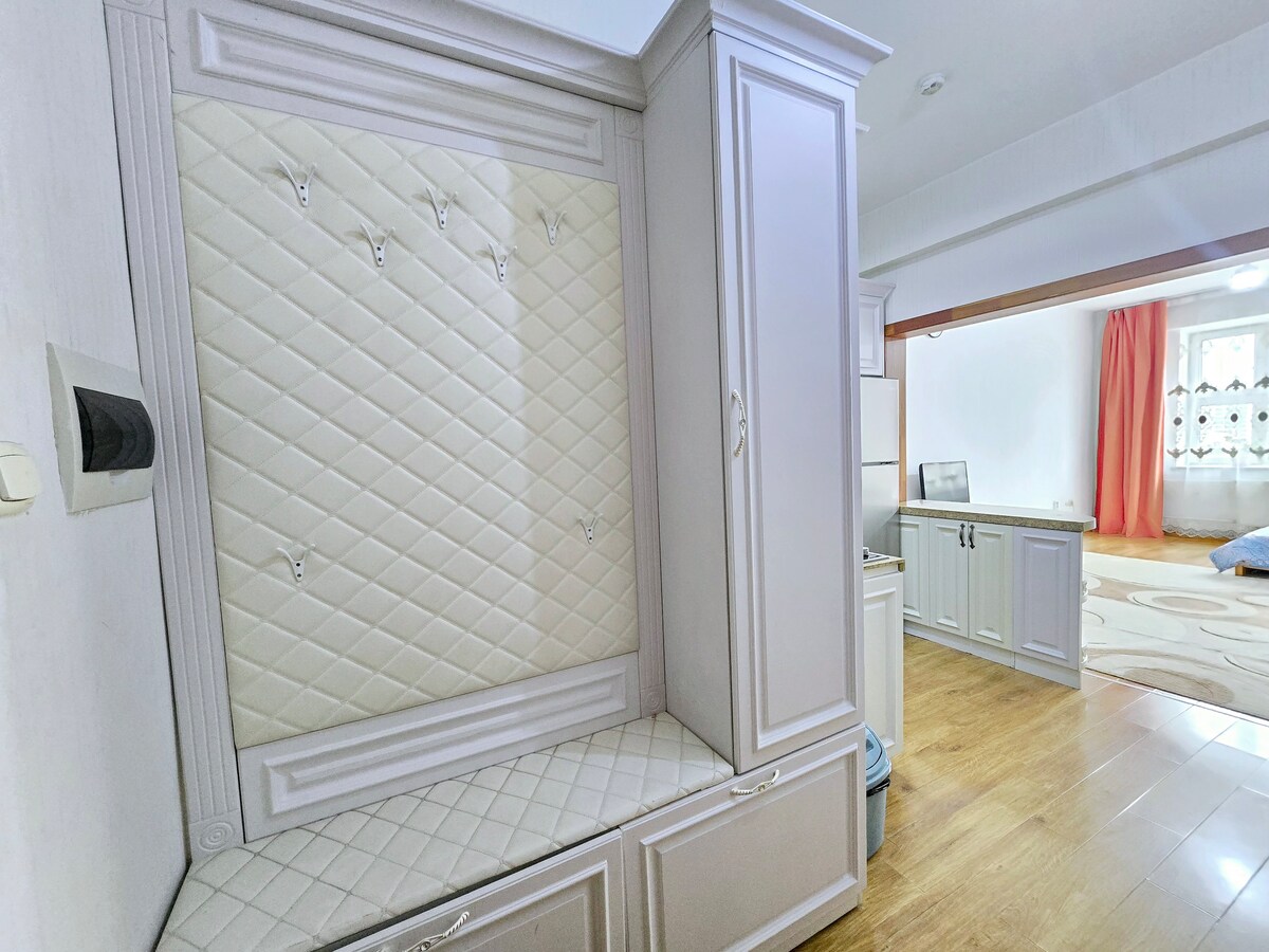 A well-designed entrance area features a quilted panel with hooks for hanging items. A built-in bench is positioned below, providing a convenient space for seating. The warm wood flooring extends into the visible living area, adding a cohesive and inviting feel.