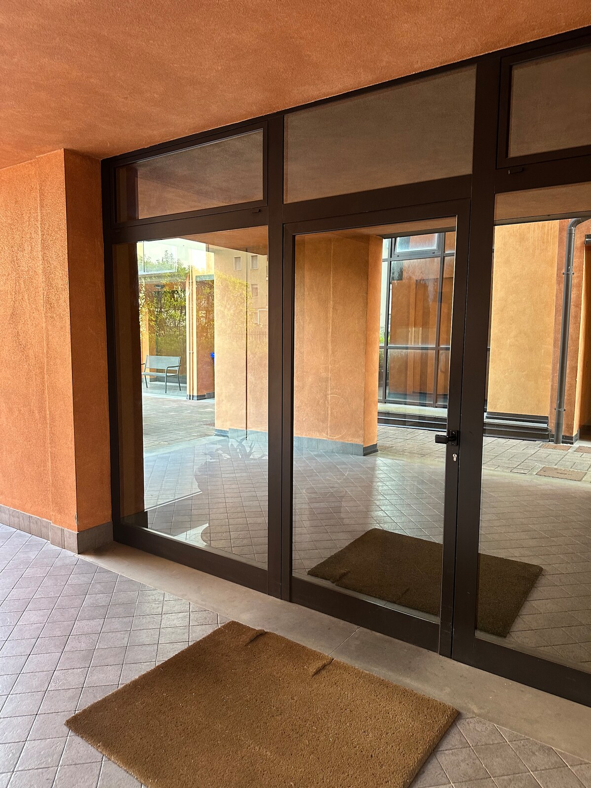 An entrance area is shown, featuring large glass doors that reflect the surroundings. The floor is finished with angled tiles, while welcome mats are positioned at the base of the doors. Natural light is observed filtering through the glass, highlighting the warm orange walls.