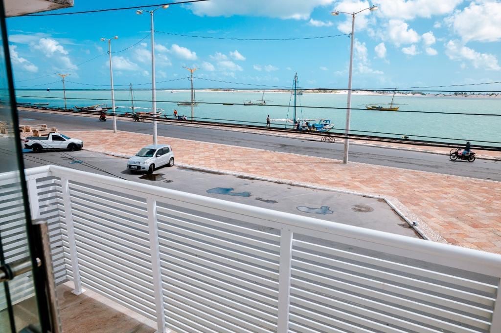 A spacious balcony offers views of the waterfront, with boats visible in the distance on serene blue waters. The area is lined with light-colored stone, and nearby vehicles are parked along the street, creating a sense of accessibility and openness.