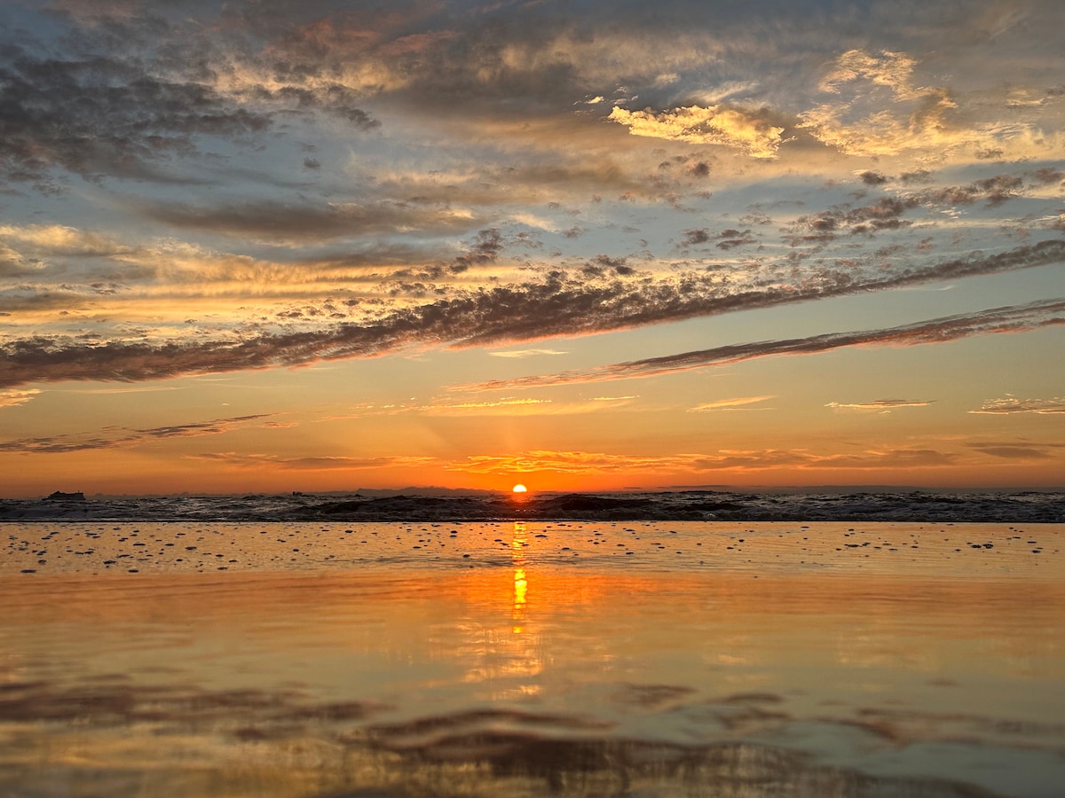 A serene sunset over the ocean is depicted, with the sun dipping below the horizon. The sky is filled with warm tones of orange and yellow, while shades of purple and blue are visible in the clouds. The water reflects the colors above, creating a calming atmosphere.