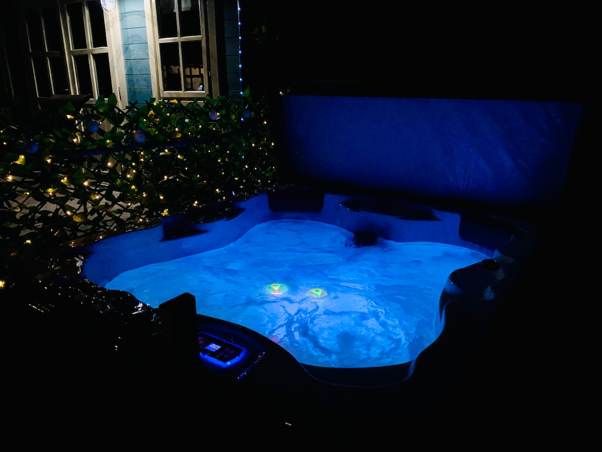 A private hot tub is illuminated with a soft blue light, surrounded by lush greenery and twinkling outdoor lights. The tub features multiple seating areas, providing a serene ambiance for relaxation.