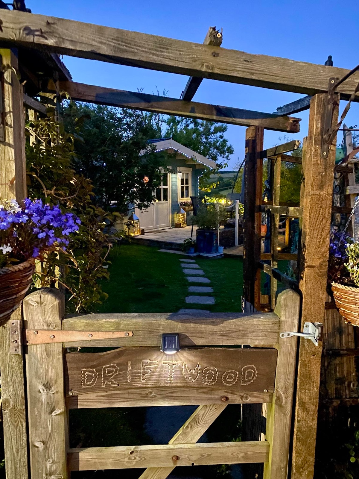 A wooden gate labeled 'DRIFTWOOD' opens to a gravel pathway, leading towards a charming cabin nestled among greenery. Flower baskets hang on either side, adding a touch of color to the serene garden setting, with soft evening light illuminating the scene.