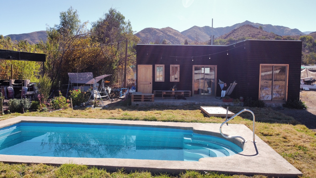 A rustic cabin is shown adjacent to a clear swimming pool, set against a backdrop of rolling hills. The cabin features wooden doors and a large window, while outdoor seating areas and lounge chairs are visible nearby, enhancing the tranquil outdoor environment.