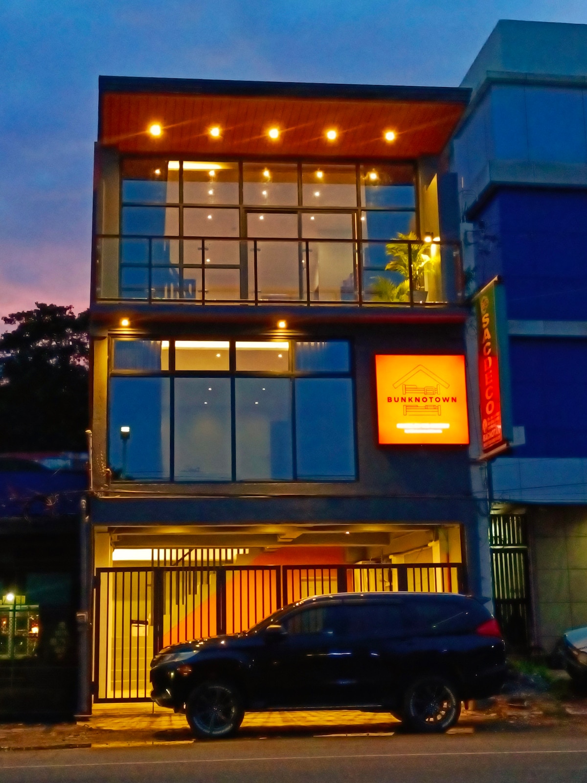 The three-story guesthouse presents a contemporary facade, featuring large glass windows that reflect the evening sky. Soft lighting highlights the entrance, while a vehicle is parked in front, indicating accessibility and convenience for guests.