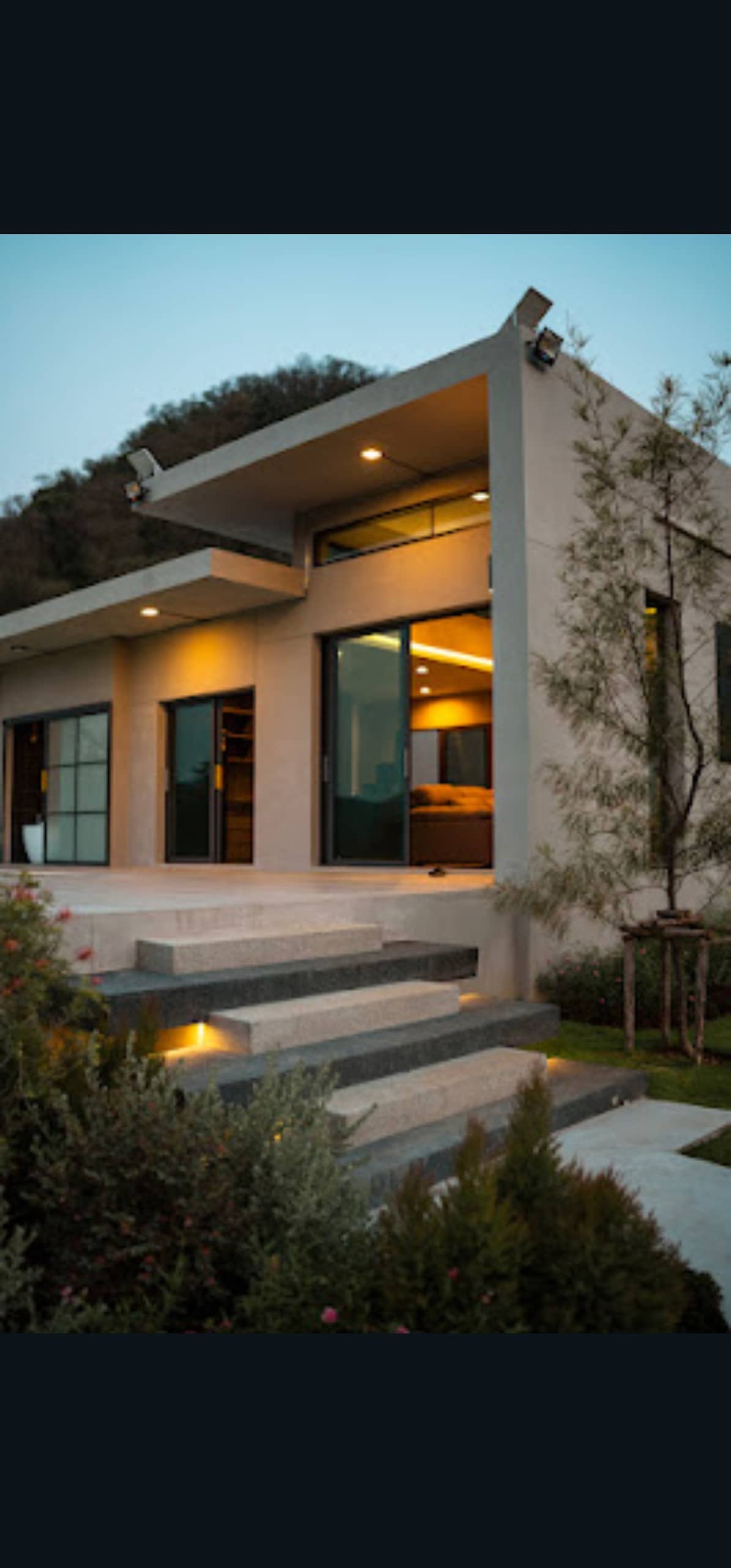 A modern villa is set against a hillside, featuring large glass doors that offer views into a well-lit interior. The entrance is framed by landscaped gardens and steps leading up to the front door, enhancing the serene ambiance of the surroundings.
