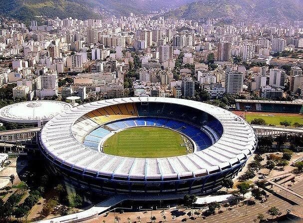 An aerial view of a prominent stadium is displayed, showcasing its circular structure with a green field at the center. Surrounding city buildings indicate an urban setting, with distant mountains adding depth to the landscape.