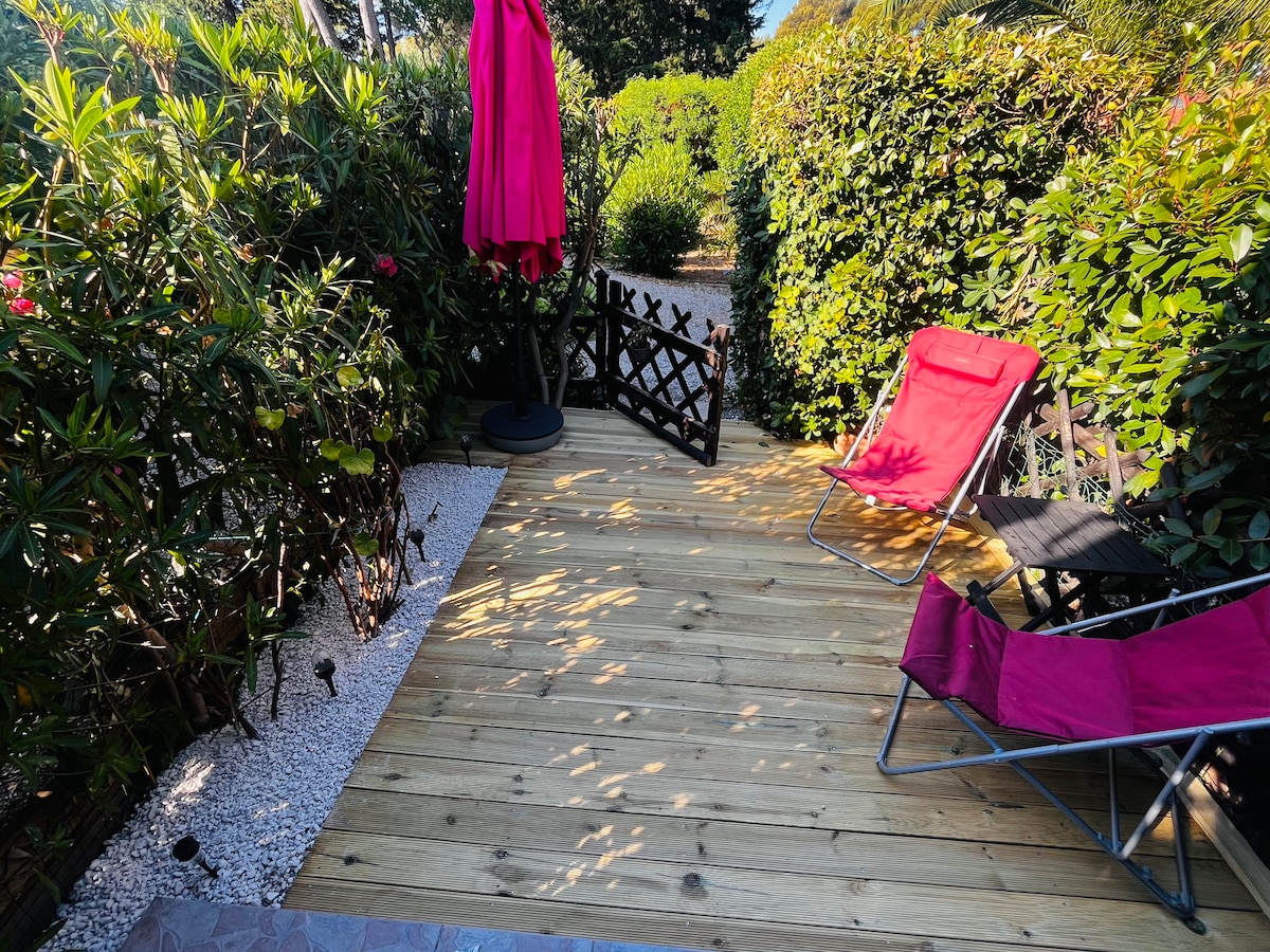 A wooden decking area is surrounded by lush greenery, providing a serene outdoor space. Two folding chairs in vibrant pink are positioned on the deck, with a pink umbrella on a stand nearby. Sunlight filters through the foliage, casting gentle shadows on the surface.