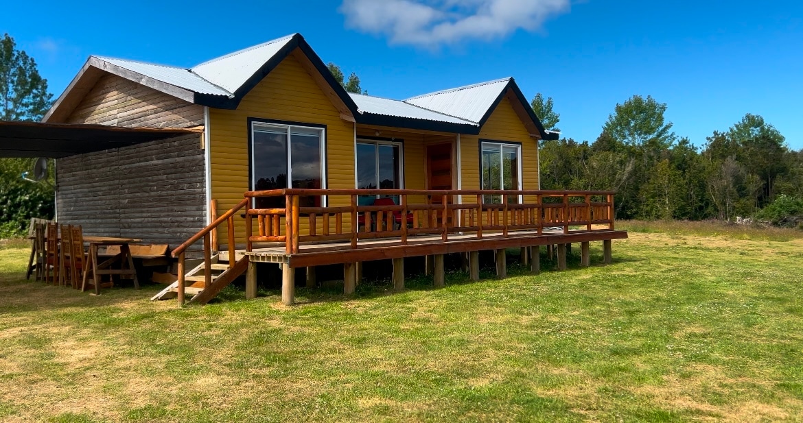 A spacious, two-story house features a bright yellow exterior with large windows inviting natural light. A wooden deck surrounds the front entrance, offering ample outdoor seating. Lush greenery and open grassland extend beyond the house, providing a serene setting.