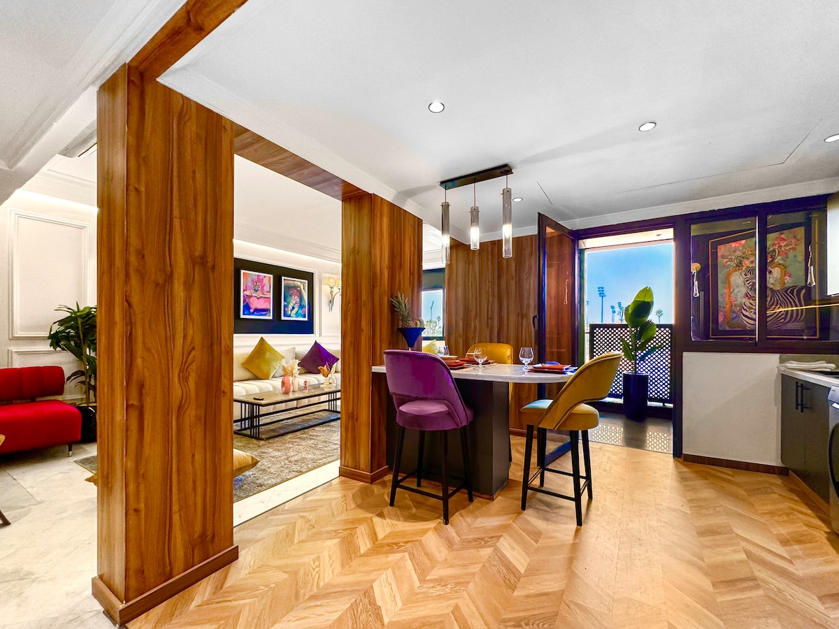 A dining area features a round table surrounded by four colorful chairs. Light wood panels create a warm ambiance, while an open space connects to a cozy living area with plush seating. Natural light filters in through the windows, enhancing the inviting atmosphere.