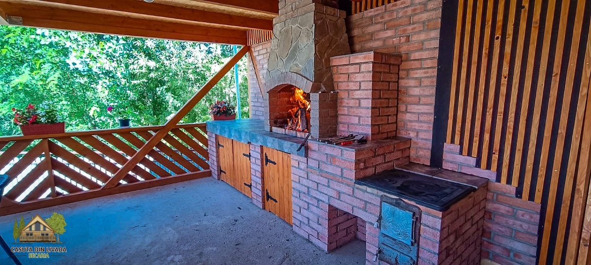 An outdoor cooking area is featured, constructed from rustic brick with a stone chimney. A barbecue grill is integrated, accompanied by a wooden countertop and cabinets. Natural light filters through the surrounding greenery, creating a serene atmosphere for meal preparation.