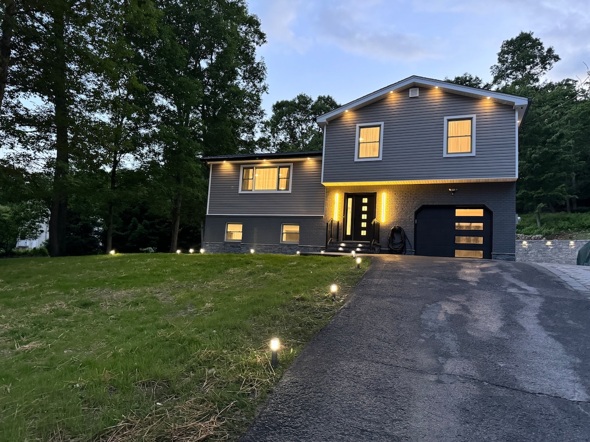 The exterior of a modern two-story home is showcased, featuring a well-manicured front lawn illuminated by soft landscape lighting. Large windows line the front, and a driveway leads to a garage door, providing ample parking space. The home is surrounded by trees, creating a serene environment.