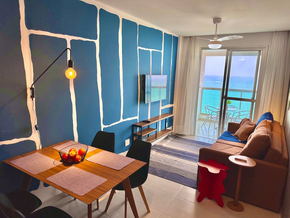 An inviting living space is depicted, featuring a brown sofa and a dining table with black chairs. A smart TV is mounted on a blue accent wall decorated with abstract patterns. Natural light streams in through the balcony door, which leads to a view of the ocean.