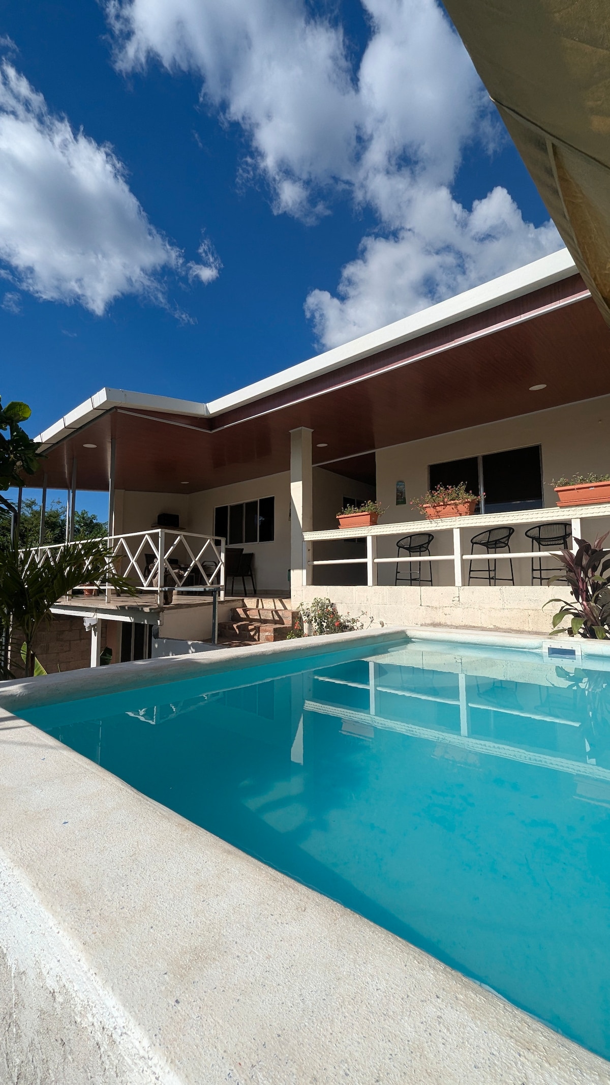 A modern house with an outdoor terrace overlooks a clear, inviting pool. The structure features large windows and a shaded patio area with seating. Surrounding greenery complements the relaxing atmosphere under a bright blue sky with scattered clouds.