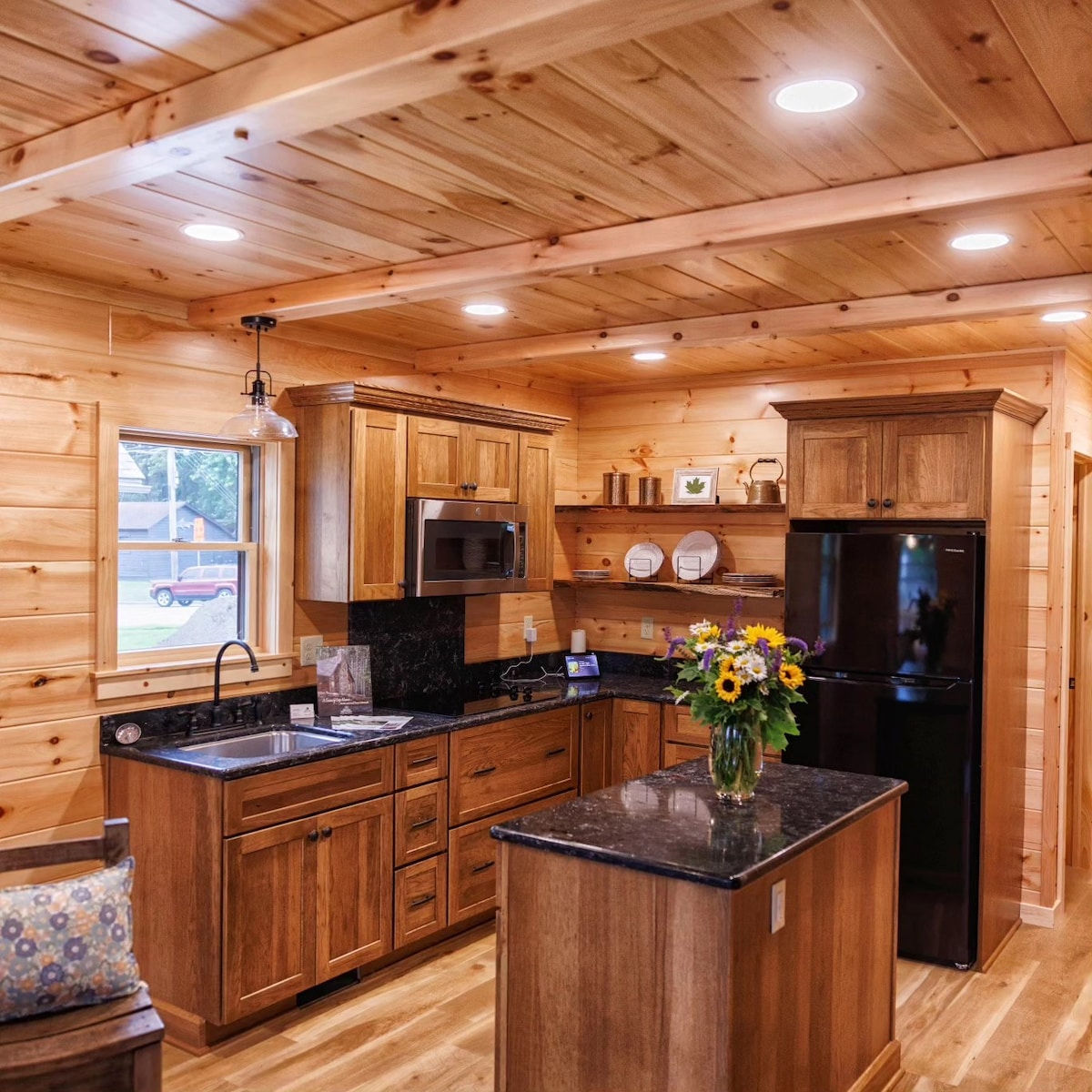 A well-equipped kitchen is presented with wooden cabinetry and granite countertops. Stainless steel appliances, including a microwave and refrigerator, are visible. A vase of sunflowers sits on the central island, providing a touch of nature. Natural light enters through a window, enhancing the warm wood tones.