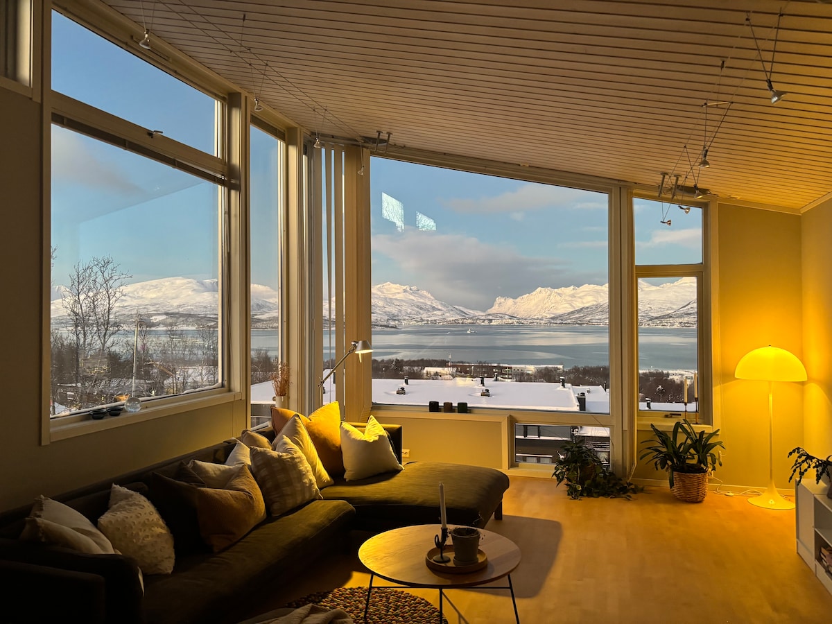A spacious living area features large windows that provide stunning views of mountains and a serene body of water. A comfortable sectional sofa, accentuated by pillows, is positioned near a warm light source, creating a cozy ambiance. Indoor plants add a touch of greenery.