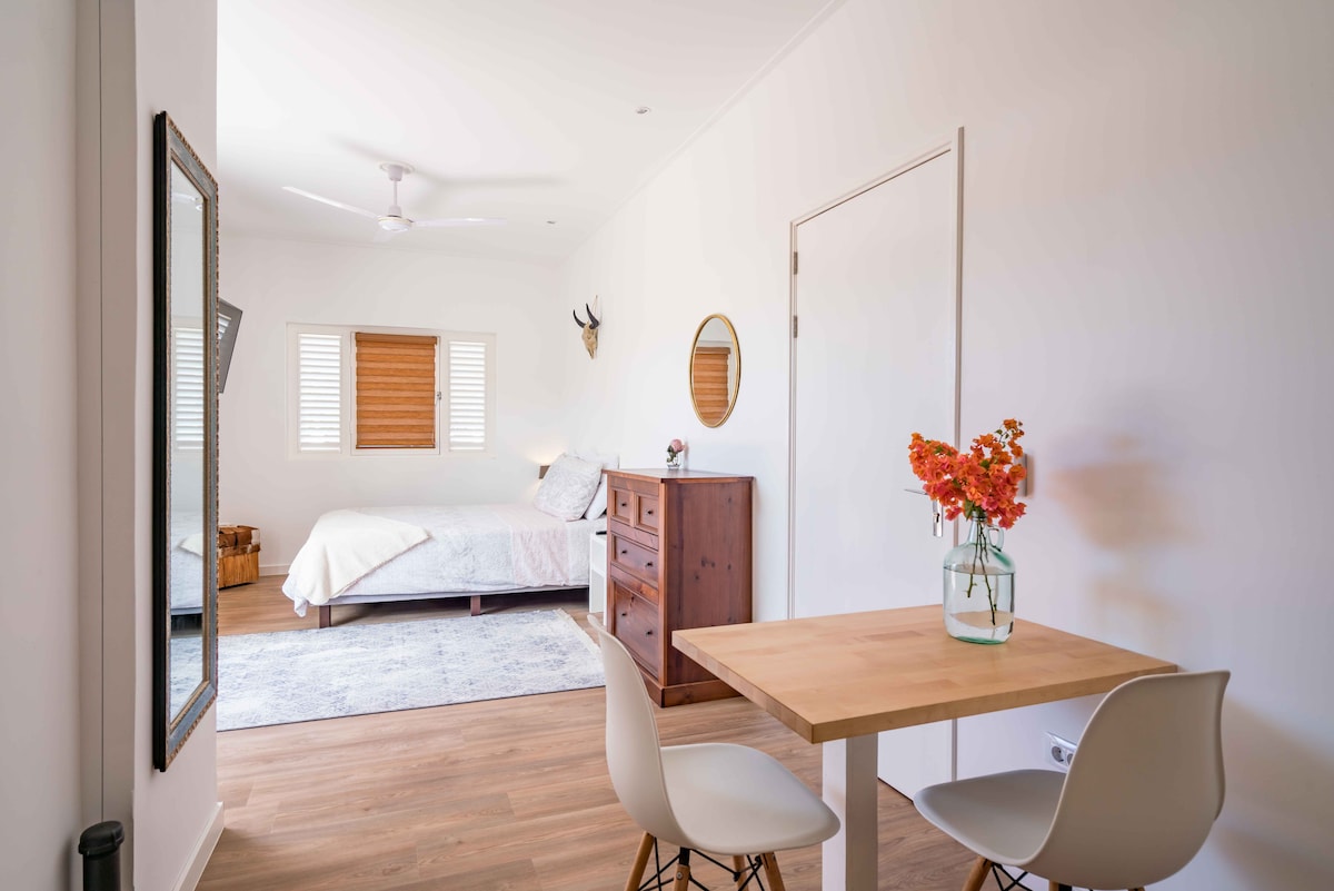 A bright and airy studio space features a spacious bed, a wooden dresser, and a small dining table with two chairs. Soft golden light enters through the window with wooden blinds, and a simple floral arrangement adds a touch of color.