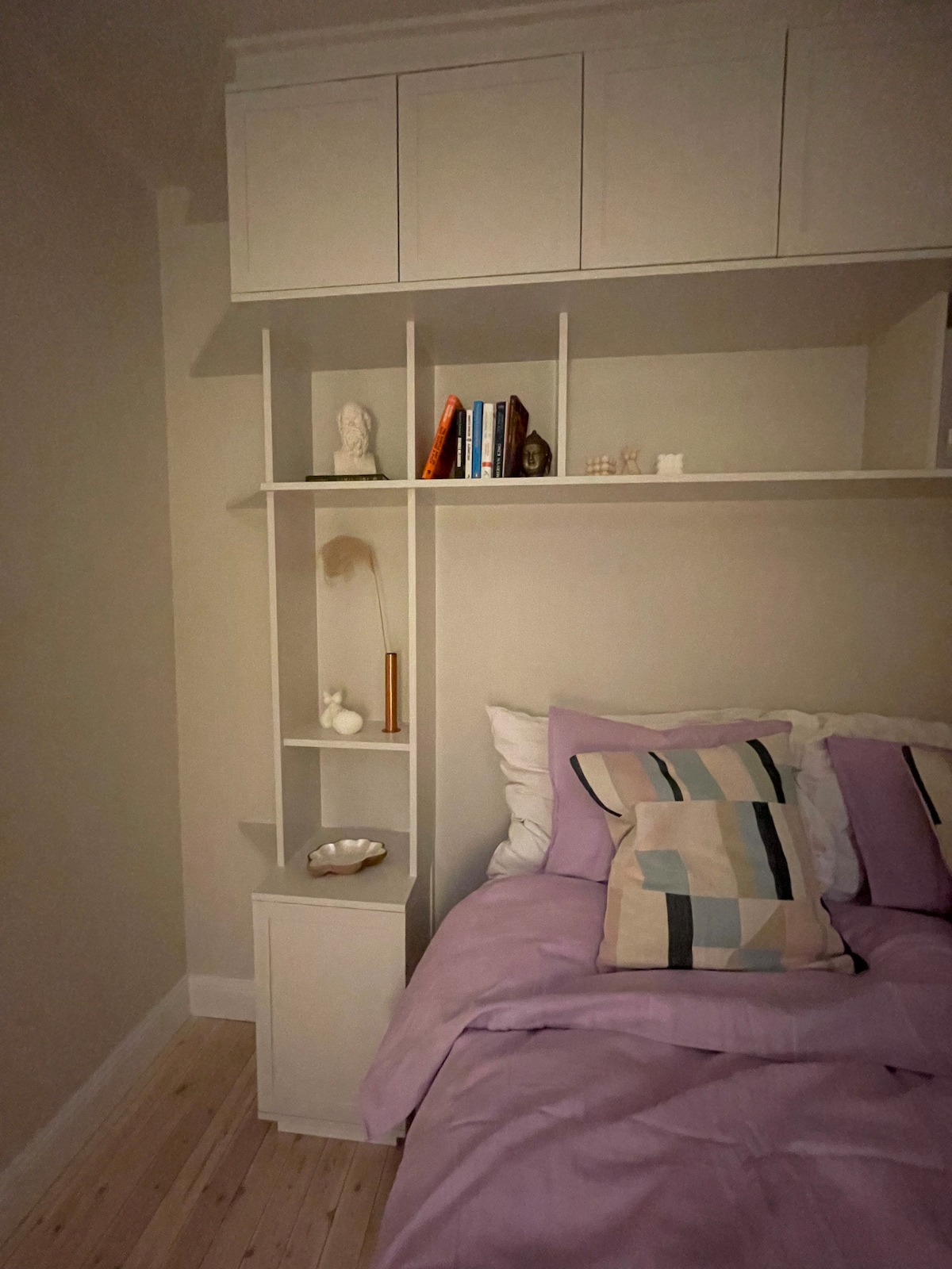 A cozy sleeping area features a spacious bed adorned with soft purple bedding. A shelving unit is positioned beside the bed, displaying decorative items and books. The warm neutral walls create a calm environment, complemented by natural light filtering in through an unseen window.