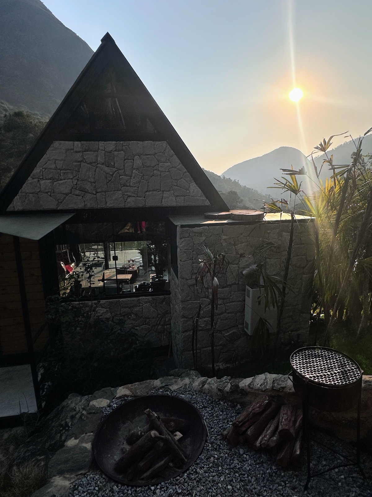 A triangular-roofed structure features stone walls and large windows offering views of rolling hills. In the foreground, firewood is arranged beside a fire pit, with sun rays illuminating the scene and creating a warm glow against the mountains in the background.