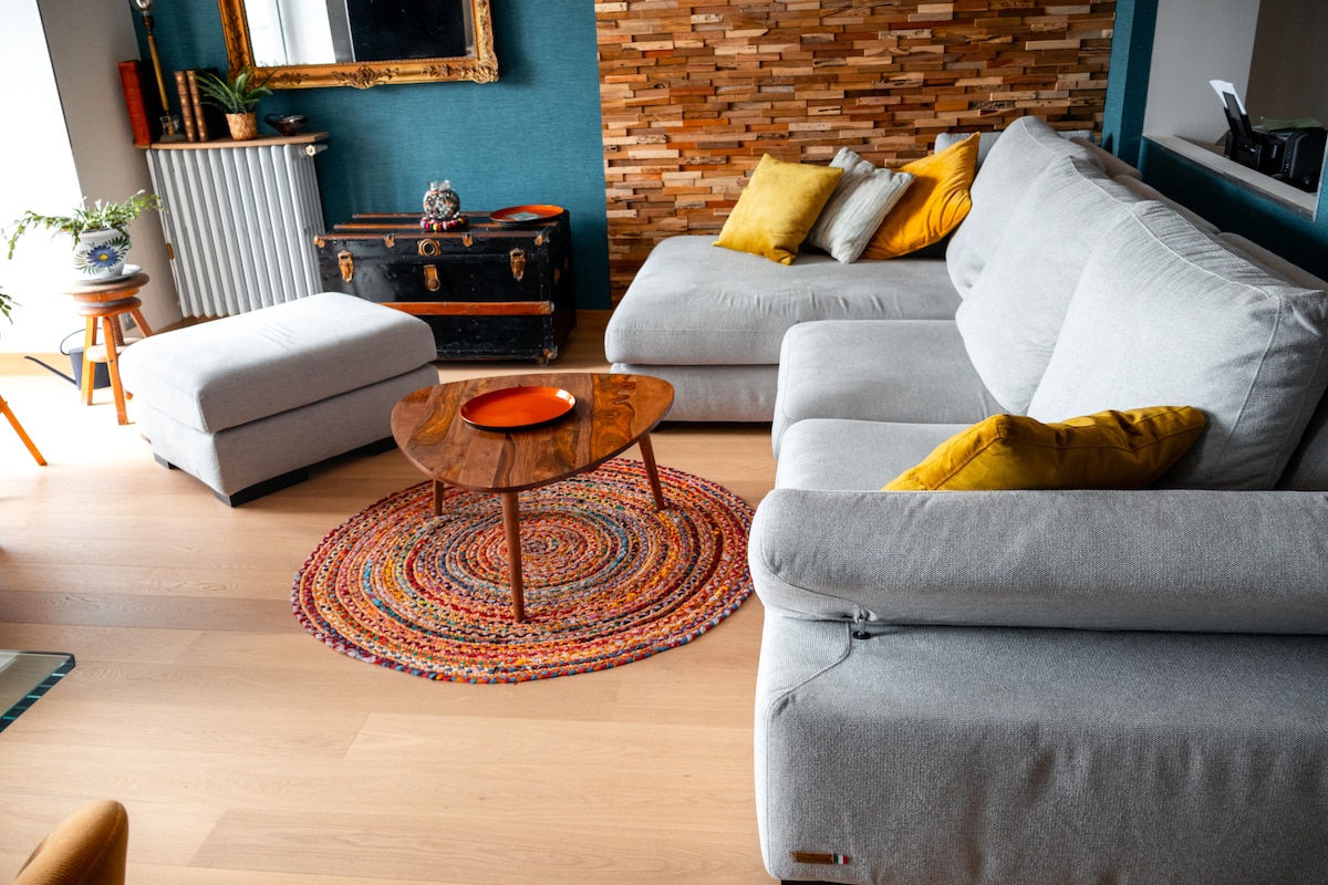 A cozy living area is presented, featuring a comfortable grey sofa with vibrant yellow cushions. A round, multicolored rug adds visual interest beneath a wooden coffee table. Natural light is welcomed through the large window, complementing a decorative wall with wooden accents.
