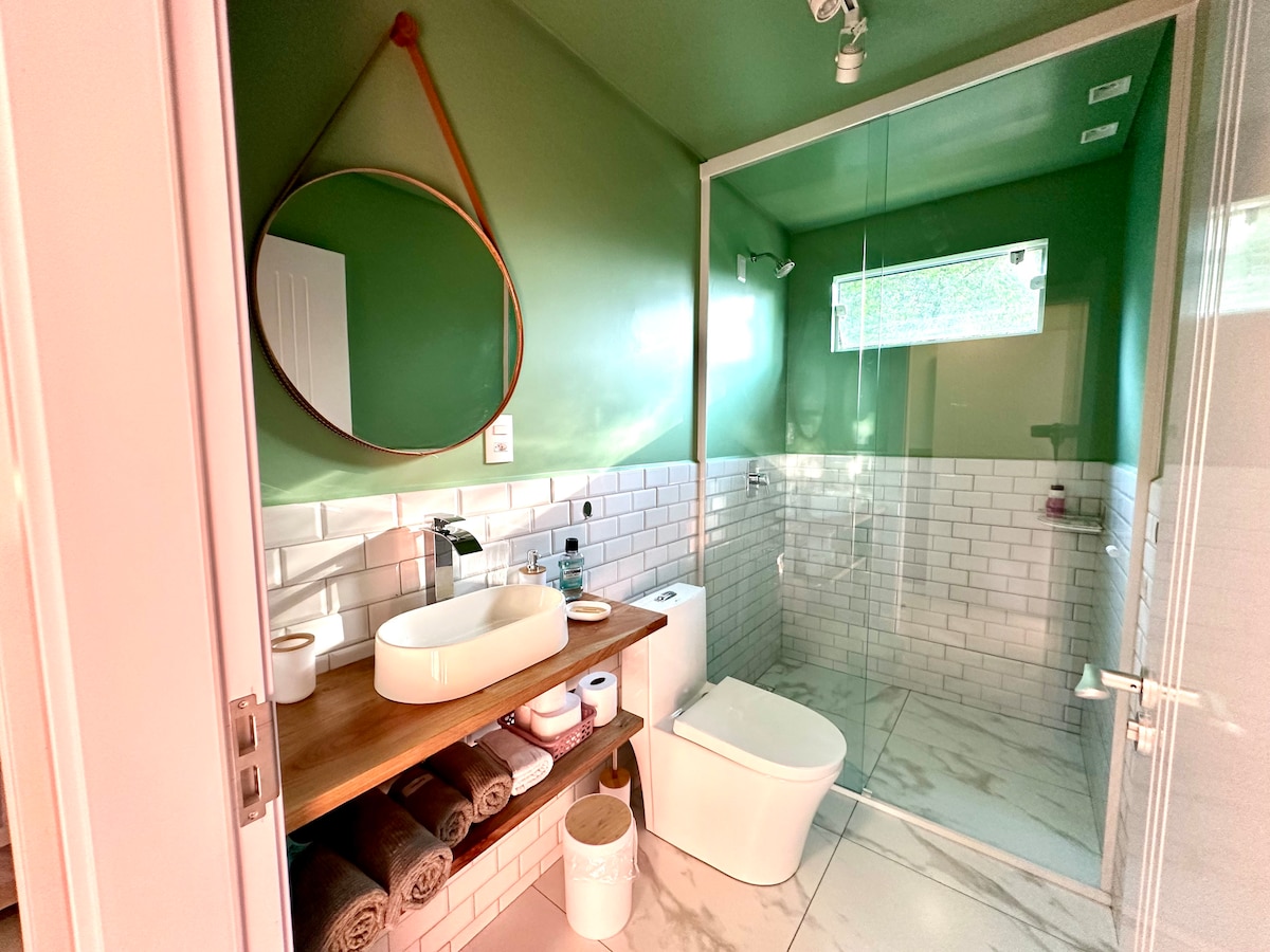 A modern bathroom is presented with green walls and natural light filtering through a window. A spacious shower area, white tiles, and a circular mirror above a wooden vanity contribute to a fresh ambience. Towels and essential toiletries are neatly arranged on the vanity.