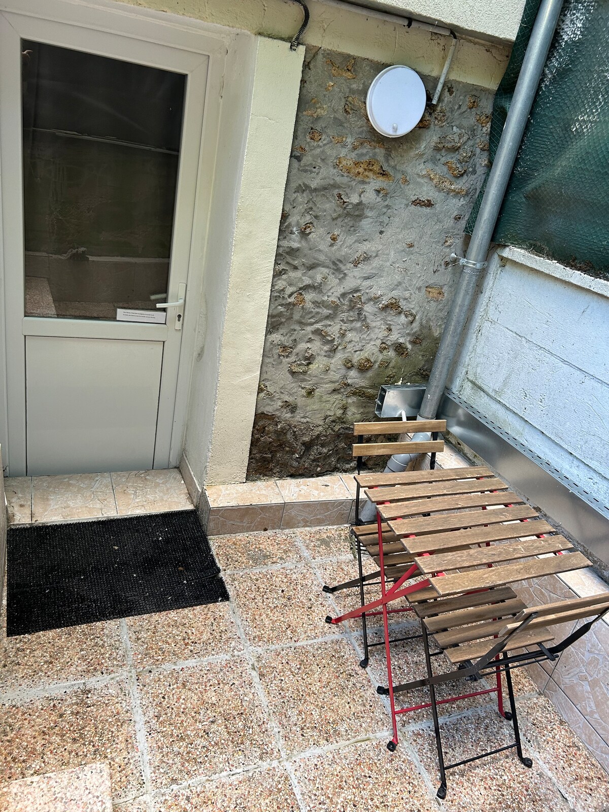 An outdoor terrace features a small wooden table and two folding chairs, set against a textured stone wall. The space is accessed through a glass-paneled door, with a rug placed at the entrance for comfort.