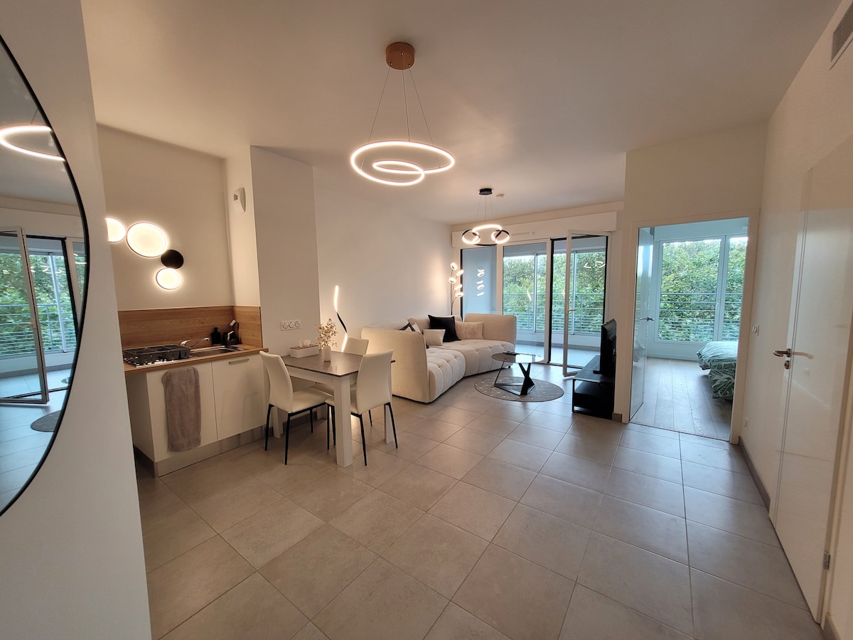 A bright living area features a comfortable sofa, armchairs, and a dining table with chairs. A modern kitchen is visible in the background, alongside large windows offering a view of greenery outside. Soft lighting fixtures add a contemporary touch to the space.