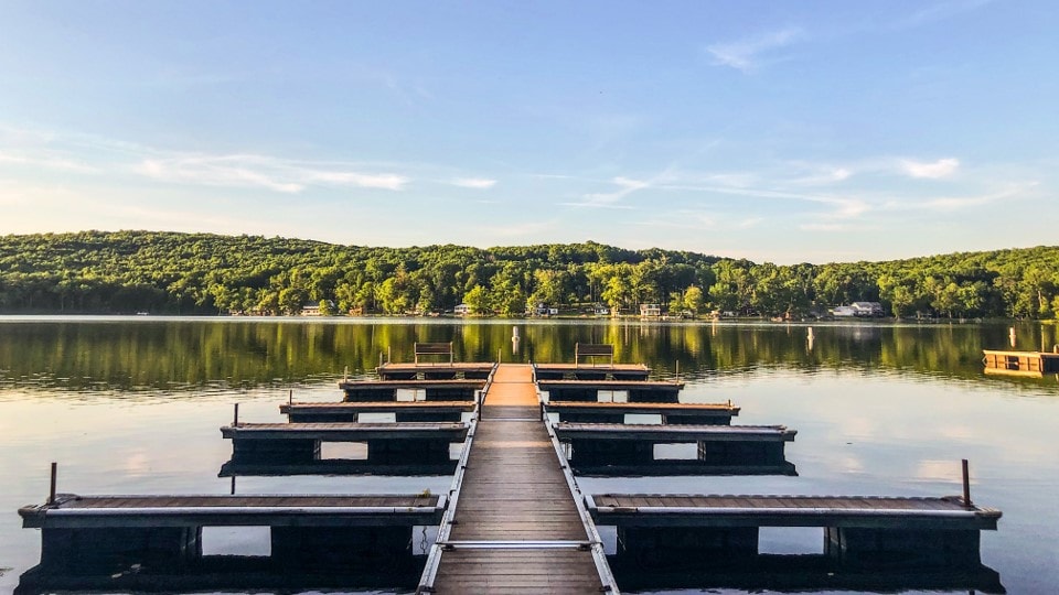 A wooden dock extends into a calm lake, flanked by several boat slips. Lush green hills surround the water, reflecting gently in the surface. Clear blue skies above complement the serene environment.