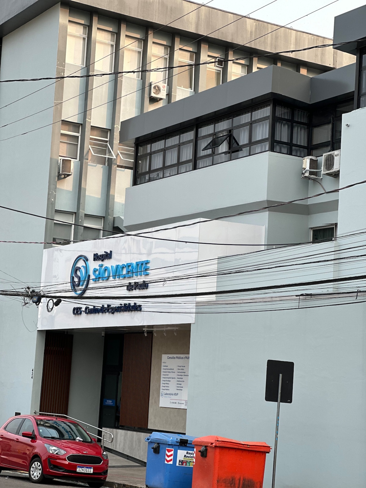 The exterior of a multi-story building is presented, featuring large windows and a modern facade. A clear sign reading 'Hospital São Vicente de Paulo' is visible. Bright blue trash bins are positioned nearby, while a red car is parked on the street in front.