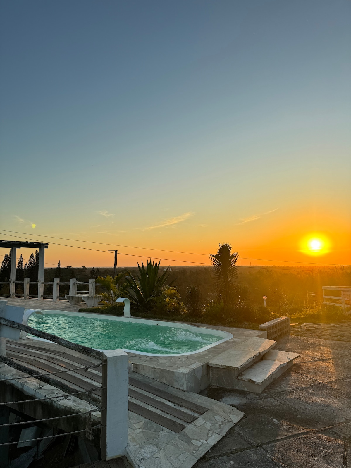 A serene outdoor area features a swimming pool surrounded by lush greenery and patio stones. The sunset casts a warm glow over the horizon, enhancing the tranquil atmosphere. The pool area is complemented by a rustic railing and distant trees, evoking a sense of calm.