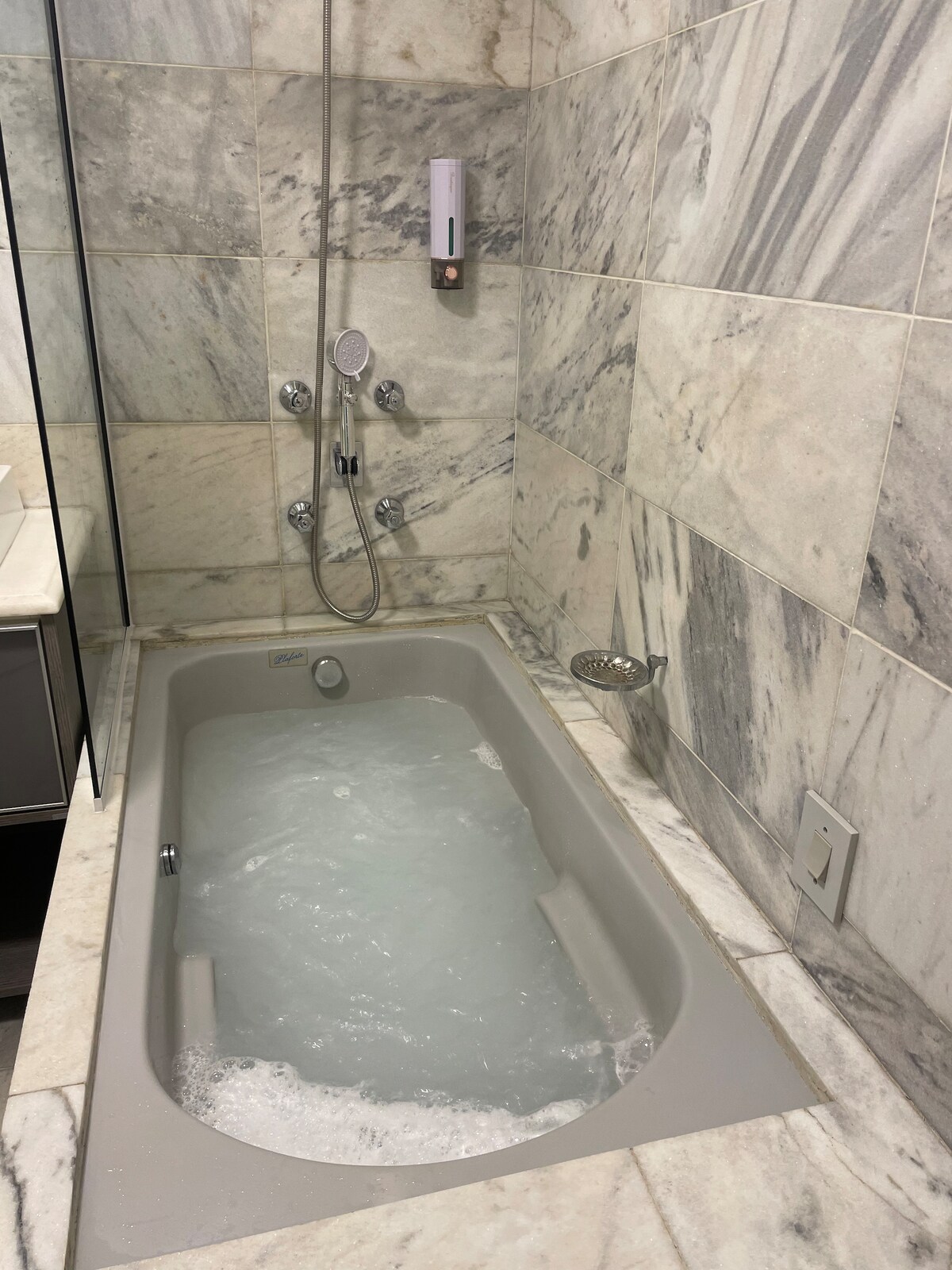 A modern bathroom features a spacious bathtub surrounded by marble walls. The bathtub is filled with bubbles, showcasing a relaxing atmosphere. Various shower fixtures are mounted on the wall, offering versatile bathing options. Natural light may stream in, enhancing the serene setting.