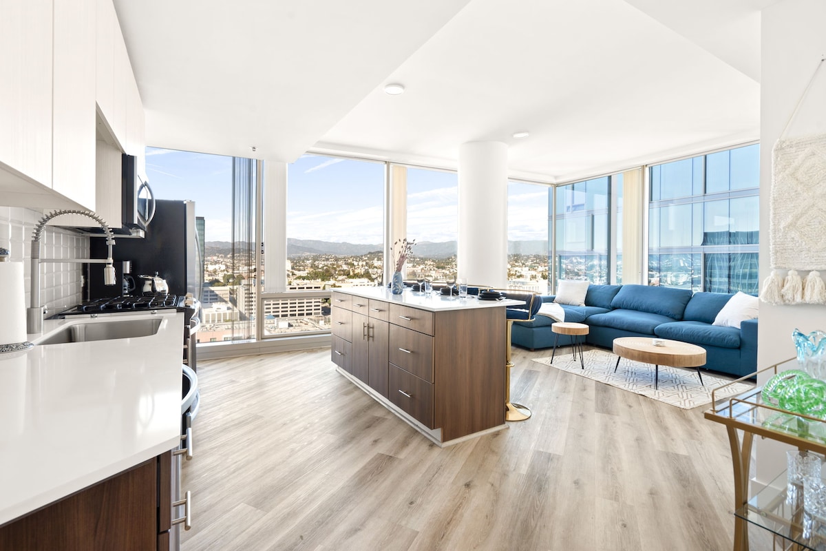 A modern kitchen with sleek cabinetry and a large island is complemented by a spacious living area featuring floor-to-ceiling windows. A comfortable blue couch and coffee tables offer ample seating, while a scenic view of the cityscape is visible beyond.