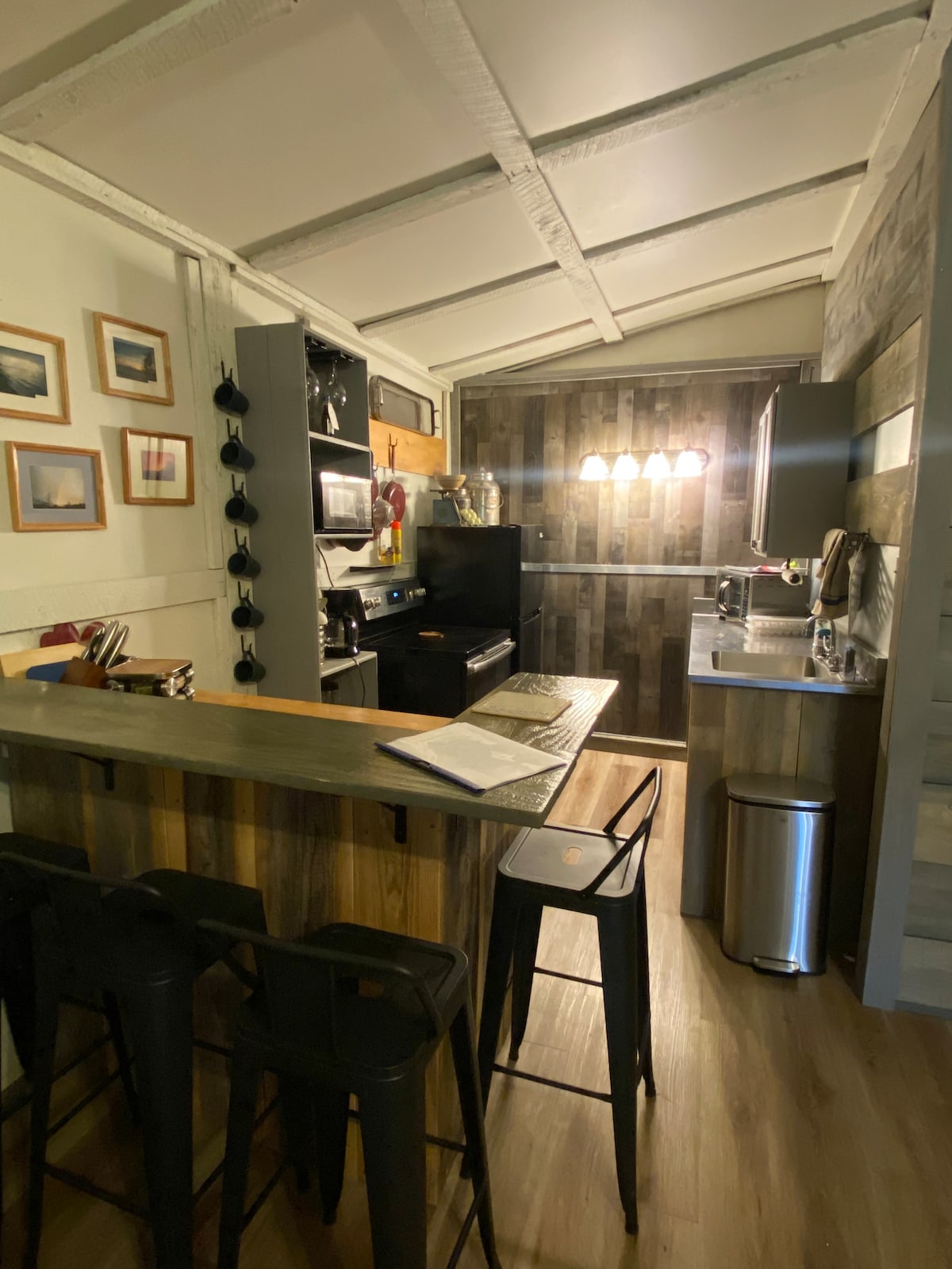 A modern kitchen is visible, featuring rustic wooden cabinetry and a bar with high stools. An electric stove is situated beside a refrigerator, and a double sink occupies the counter space. Soft lighting enhances the warm ambiance, while framed artwork adorns the walls.