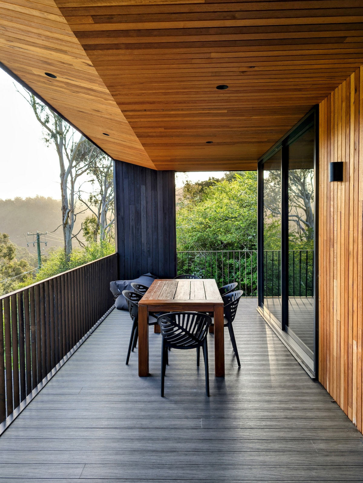 A covered deck is featured, showcasing a wooden dining table surrounded by four black chairs. The space is framed by wooden paneling and has expansive glass doors that open to the lush greenery beyond, offering a peaceful outdoor retreat.