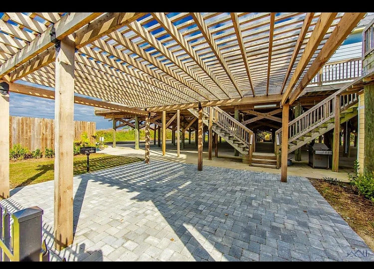 A spacious outdoor area is featured with a wooden pergola, providing shade. The ground is paved with stone, creating a smooth surface. Stairs lead up to the condo entrance, while a grilling area is situated nearby, surrounded by well-maintained landscaping.