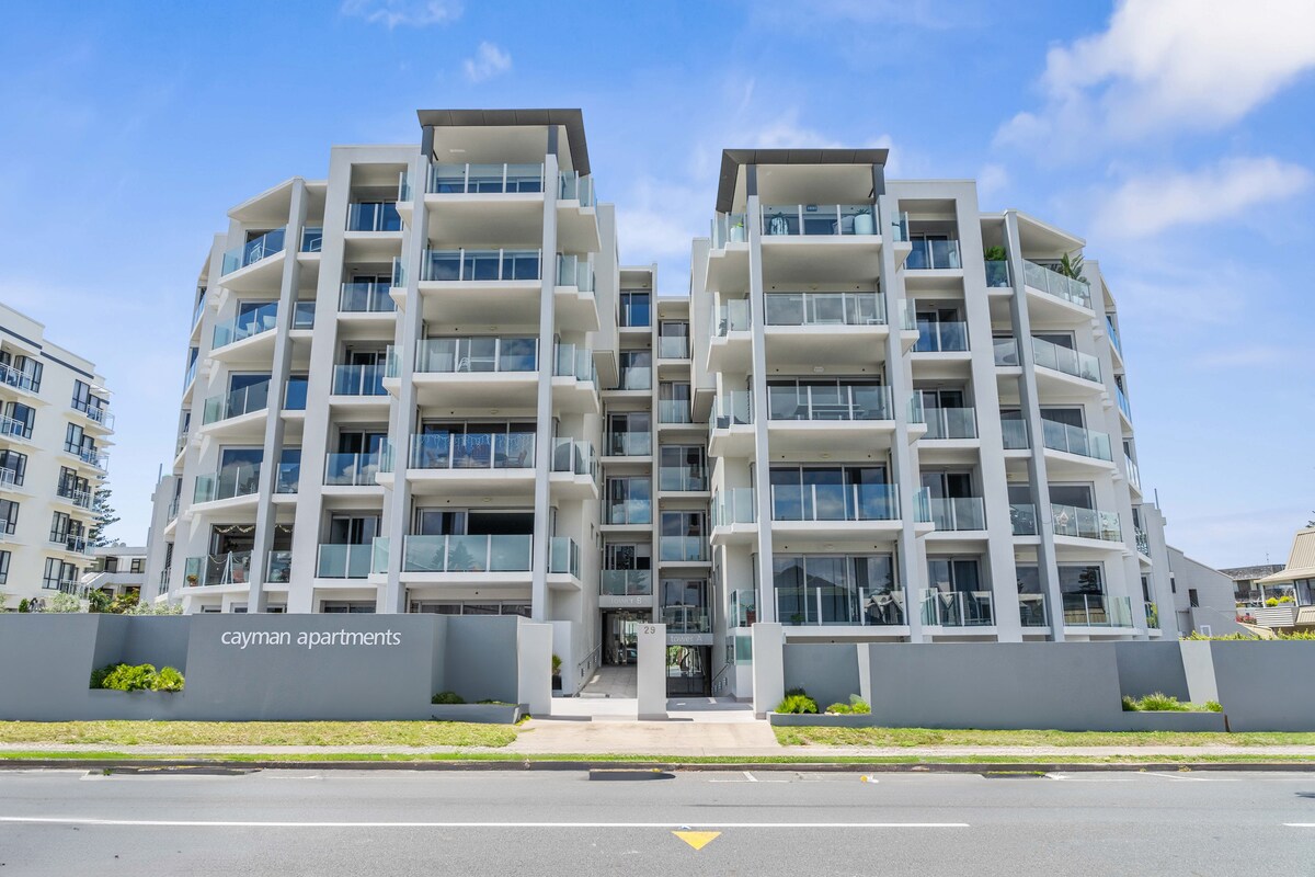 A modern apartment building is shown, featuring a contemporary design with multiple balconies. The façade is light-coloured, complemented by glass railings. The landscaping includes simple greenery near the entrance, while the street is visible in the foreground.