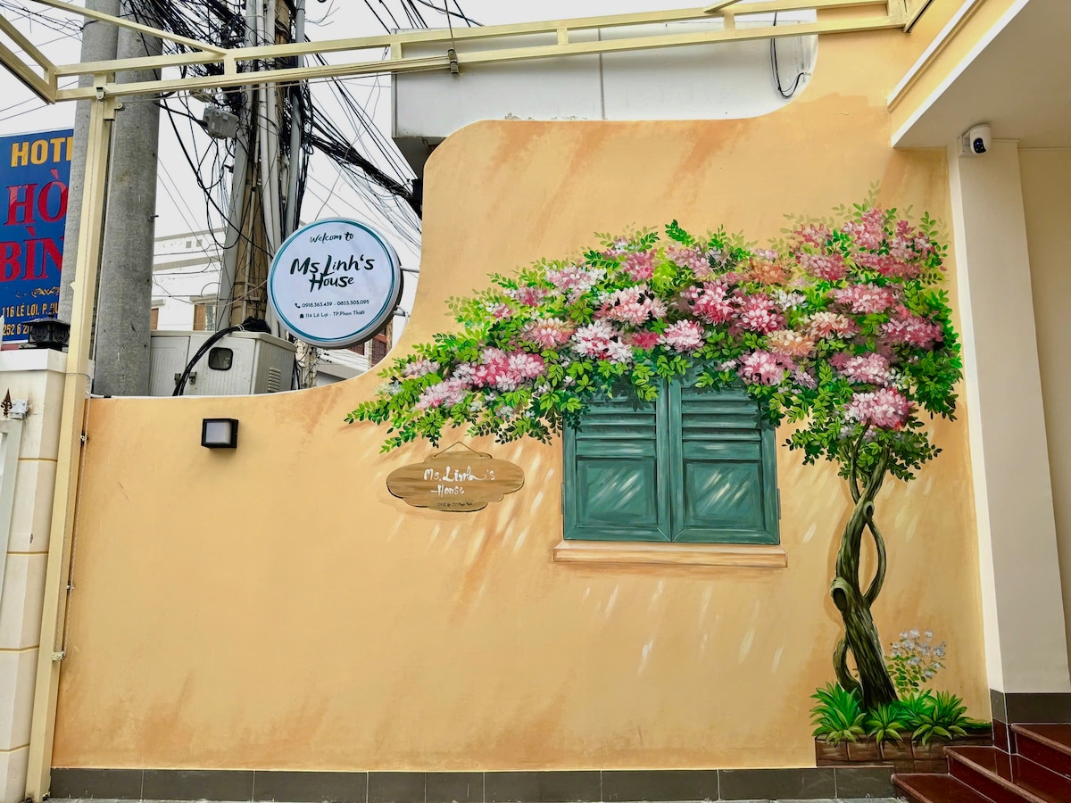 A soft peach-colored wall features a decorative mural of blooming flowers and a window with green shutters. A sign displaying 'Ms Linh’s House' is prominently placed above, while visible utility lines add a context of urban life.