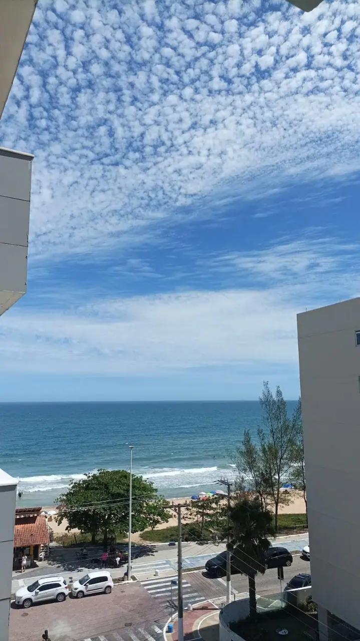 A view from the sixth floor showcases a blue sky adorned with wispy clouds above the ocean. The water gently meets the sandy shore, with some greenery and beach umbrellas visible below, highlighting the coastal atmosphere.