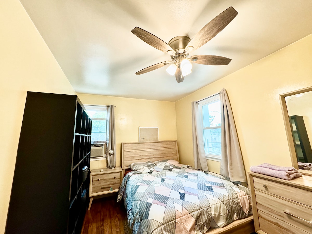 A neatly arranged bedroom features a spacious bed covered with a patterned quilt. A ceiling fan provides airflow while a window allows natural light to brighten the room. Dark wood furniture, including a dresser and shelving unit, complements the light walls and adds utility.