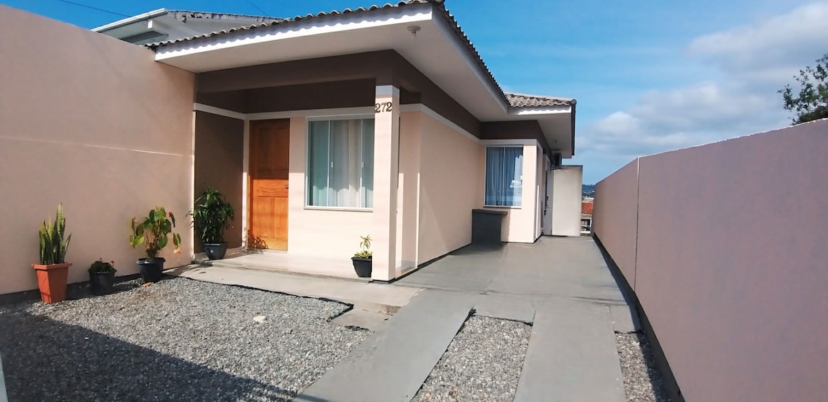 A single-story home is showcased, featuring a wooden front door and large windows framed by light-colored walls. The entrance is bordered by potted plants and a gravel pathway, leading to an outdoor area that appears spacious and welcoming.