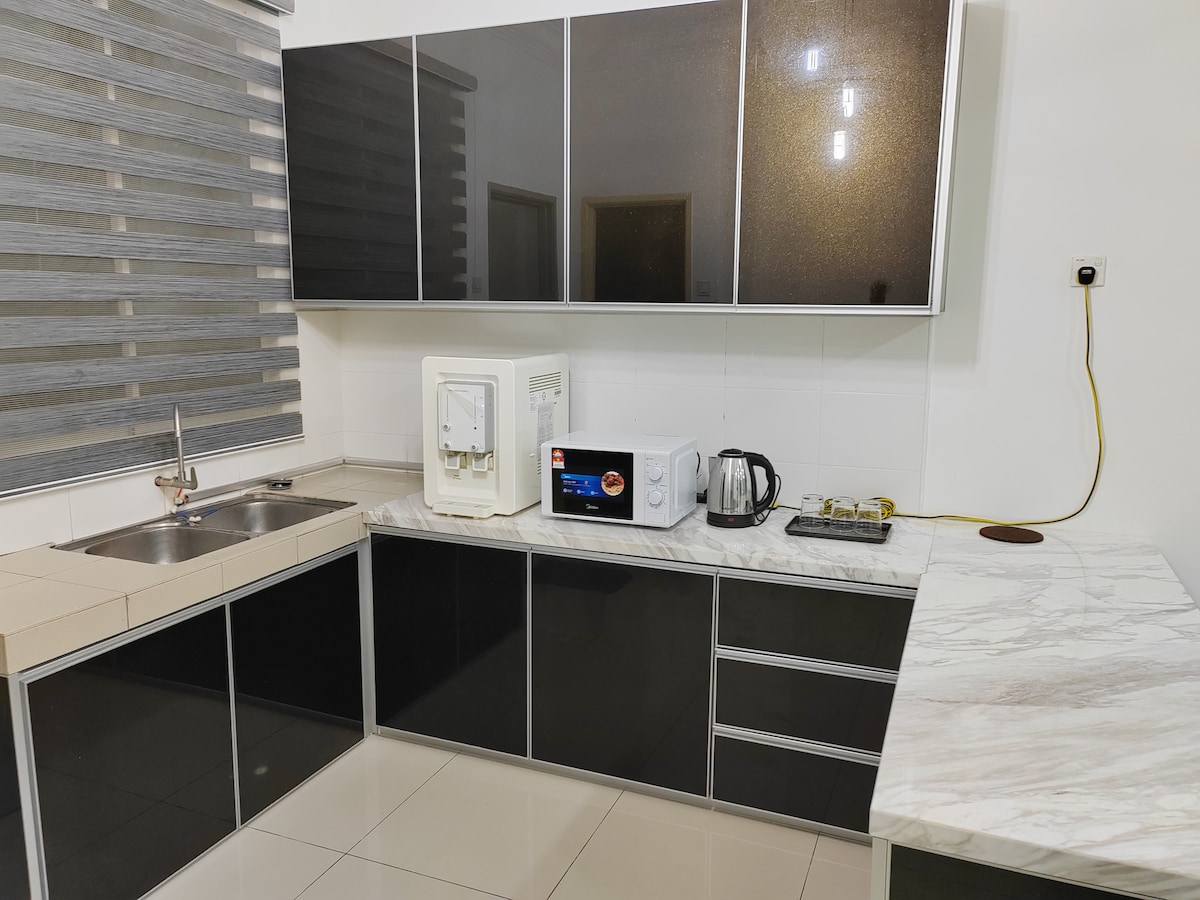 A modern kitchen area showcases sleek black cabinetry paired with a marble countertop. Essential appliances include a refrigerator, microwave, and electric kettle. A stainless steel sink is positioned near a window with layered blinds, providing a functional and stylish cooking environment.