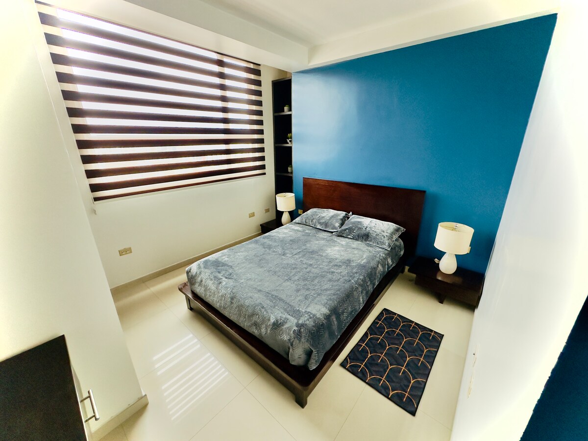 A spacious bedroom features a dark wood bed with a comfortable gray bedspread. Two bedside lamps provide soft lighting, and a colorful area rug lays on the polished floor. A window with striped shades allows natural light to filter in, illuminating the vibrant blue accent wall.