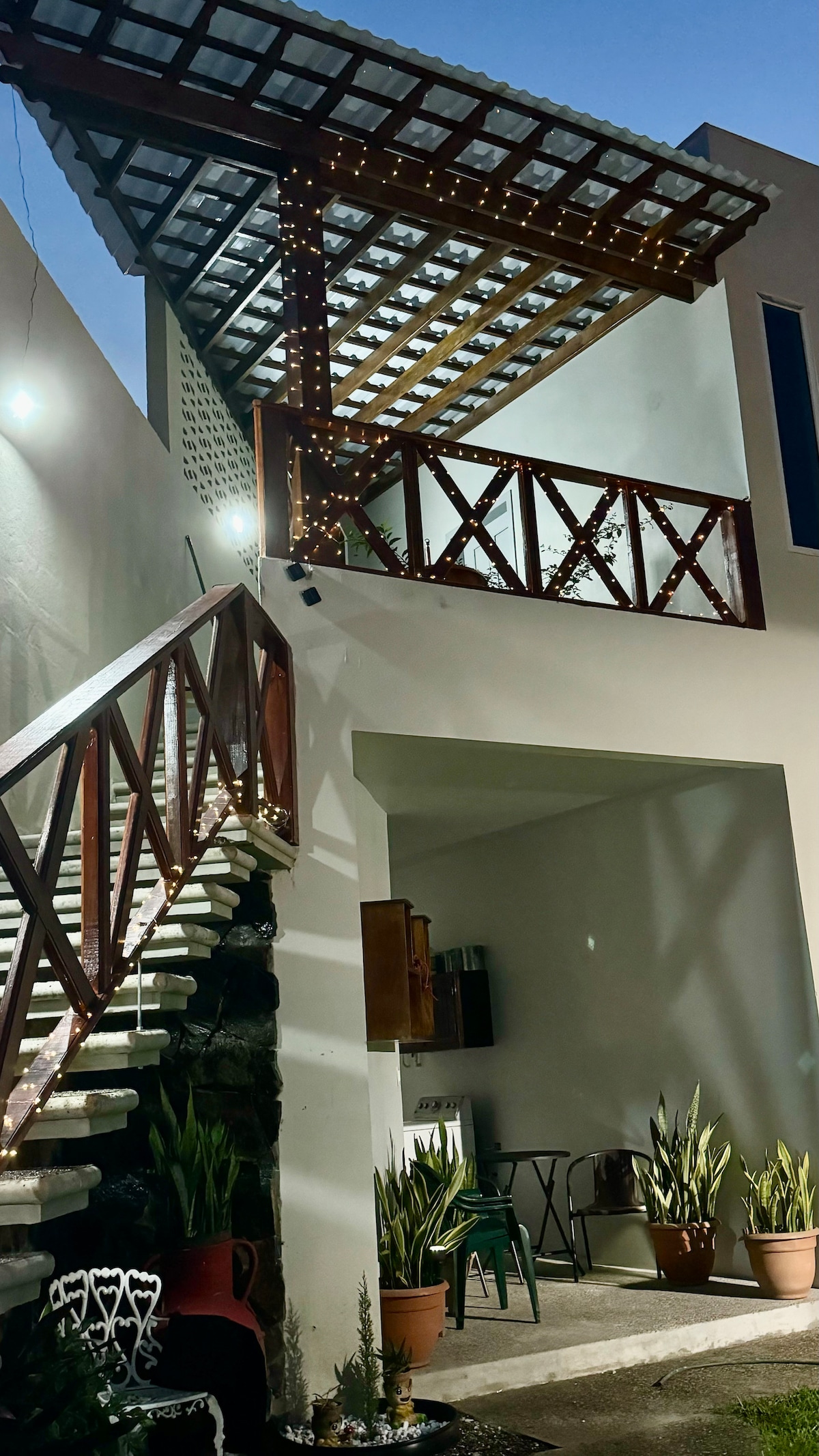 An outdoor area is presented, featuring a staircase leading to a balcony with wooden railings adorned with lights. Potted plants line the space, and a small table and chairs are visible in the shadows, creating a cozy atmosphere for relaxation.