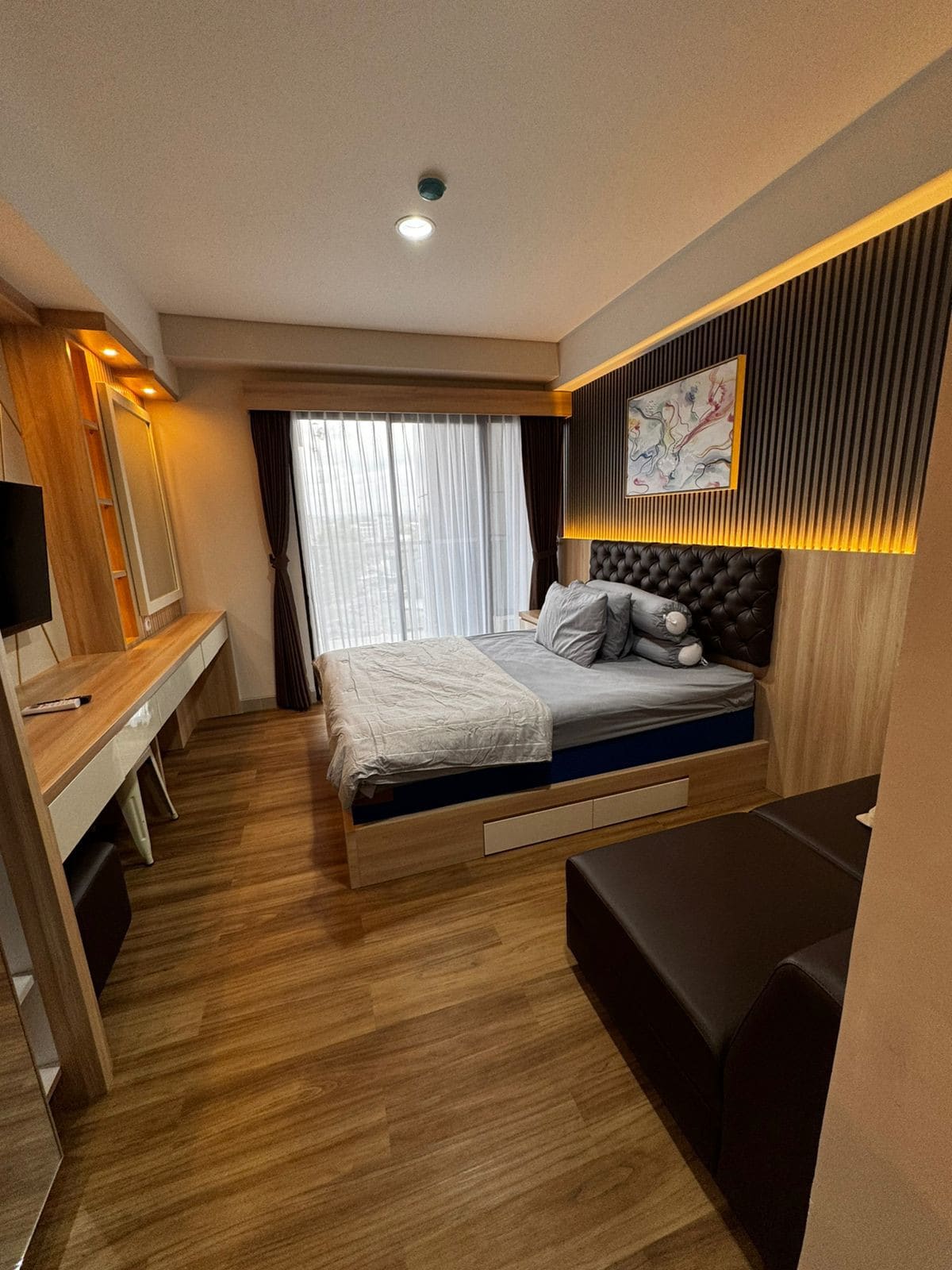 A minimalist studio apartment features a queen-size bed with a tufted headboard, accompanied by bedside decor. A spacious window allows natural light to fill the room, with sheer curtains partially drawn. A modern workstation and a cozy seating area are also visible.