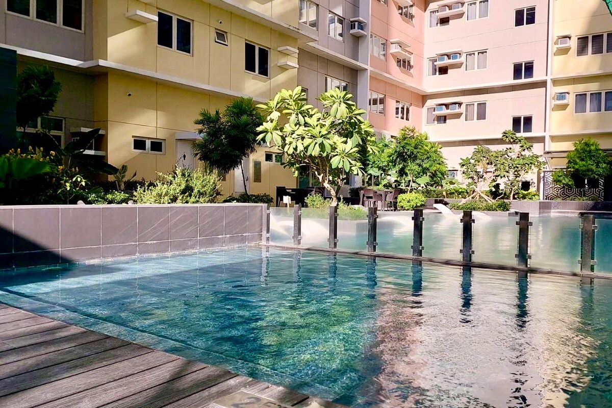 A serene pool area is visible, bordered by lush greenery and well-maintained landscaping. The shimmering water reflects the surrounding buildings and calming environment, while wooden decking provides a relaxing space for guests. Sunlight filters through the trees, enhancing the tranquil setting.