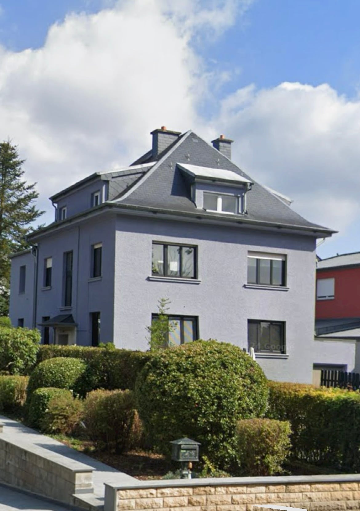 A multi-story house with a gray exterior is framed by neatly trimmed bushes and trees. Large windows are visible on each level, allowing natural light to enter. The roof features multiple peaks, and a clear blue sky is visible above.