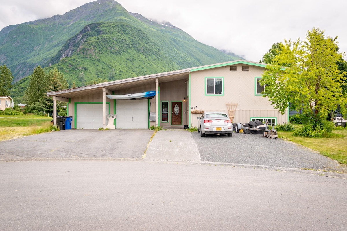A spacious single-story house is nestled in a scenic landscape, featuring a large front yard with grass and trees. The structure has a modern design with a wide driveway and parking area, complemented by a mountain backdrop and cloudy skies overhead.