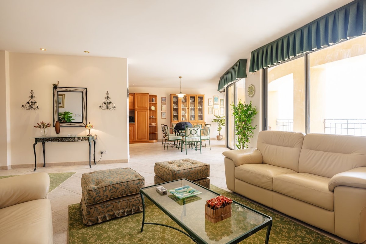 A spacious living area features light-colored sofas and a decorative glass coffee table. Natural light fills the room through large windows. A dining area is visible in the background, surrounded by elegant furniture and green plants.