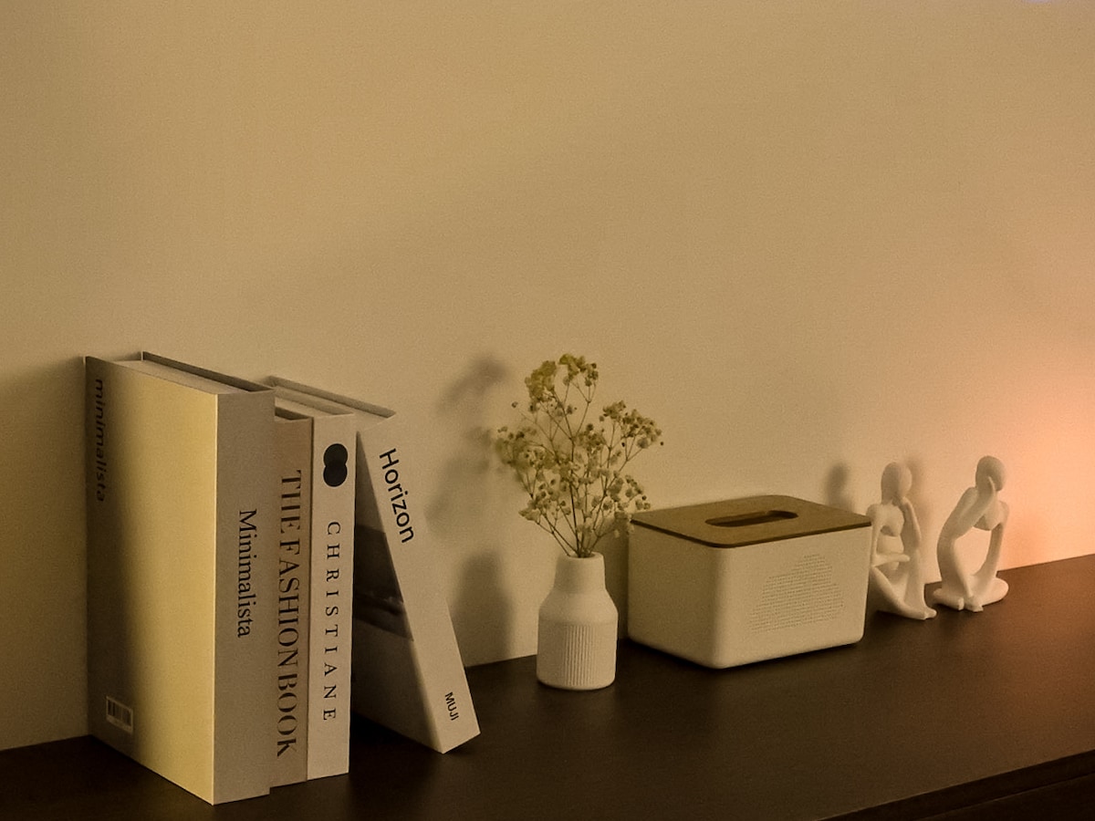 A simple bookshelf displays three books, including 'The Fashion Book.' A small vase holds delicate flowers, while a light-colored container is positioned next to a pair of decorative figurines. The warm glow from a nearby light enhances the subtle décor.