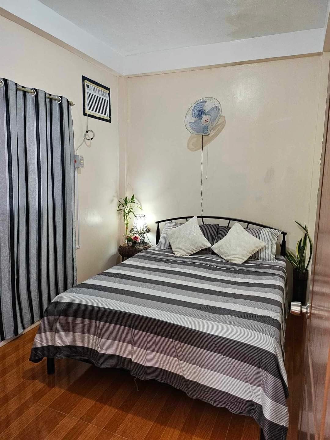 A cozy bedroom features a striped bedspread over a spacious bed, with two decorative pillows placed at the head. A small plant adds a touch of greenery beside a bedside table. Vertical striped curtains frame a window, complemented by a ceiling fan mounted above.