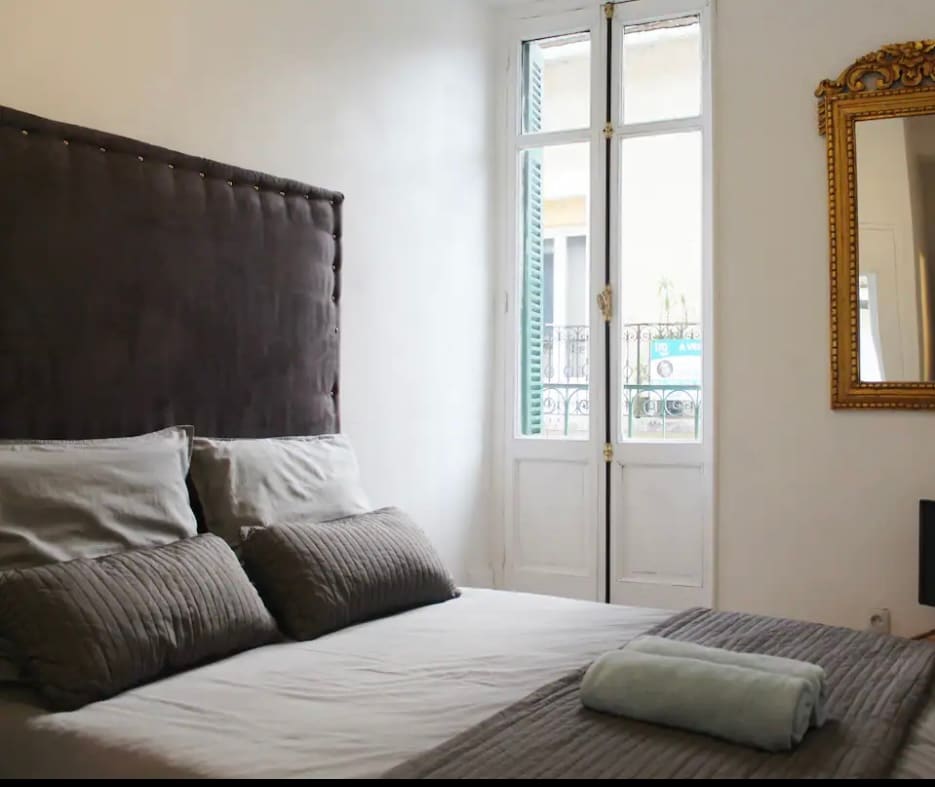 A serene bedroom is featured, with a spacious bed adorned with textured gray linens and plush pillows. Natural light enters through the window, illuminating the decor, while a large ornate mirror is positioned to reflect the room's calming ambiance.